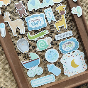 Stickers naissance scrapbooking posés sur une toile de jute : landau, lune étoilée et étiquettes de bienvenue